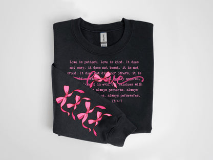 May include: Black sweatshirt with pink text and bow accents. The text reads "Love is patient, love is kind..." with the word "Love" in a cursive font. Pink bows are on the left sleeve. The sweatshirt is folded.