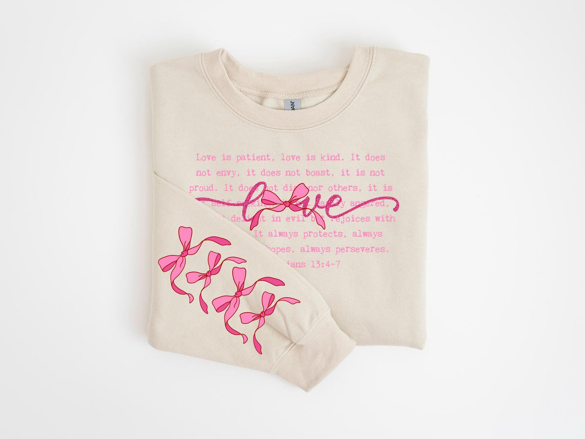 May include: A beige sweatshirt with the word "love" in pink script and the Bible verse, 1 Corinthians 13:4-7, printed in pink. The sleeve features pink bow graphics.