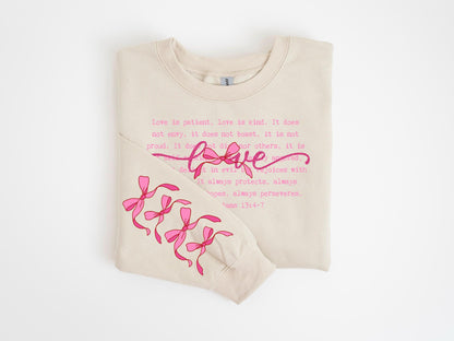 May include: A beige sweatshirt with the word "love" in pink script and the Bible verse, 1 Corinthians 13:4-7, printed in pink. The sleeve features pink bow graphics.