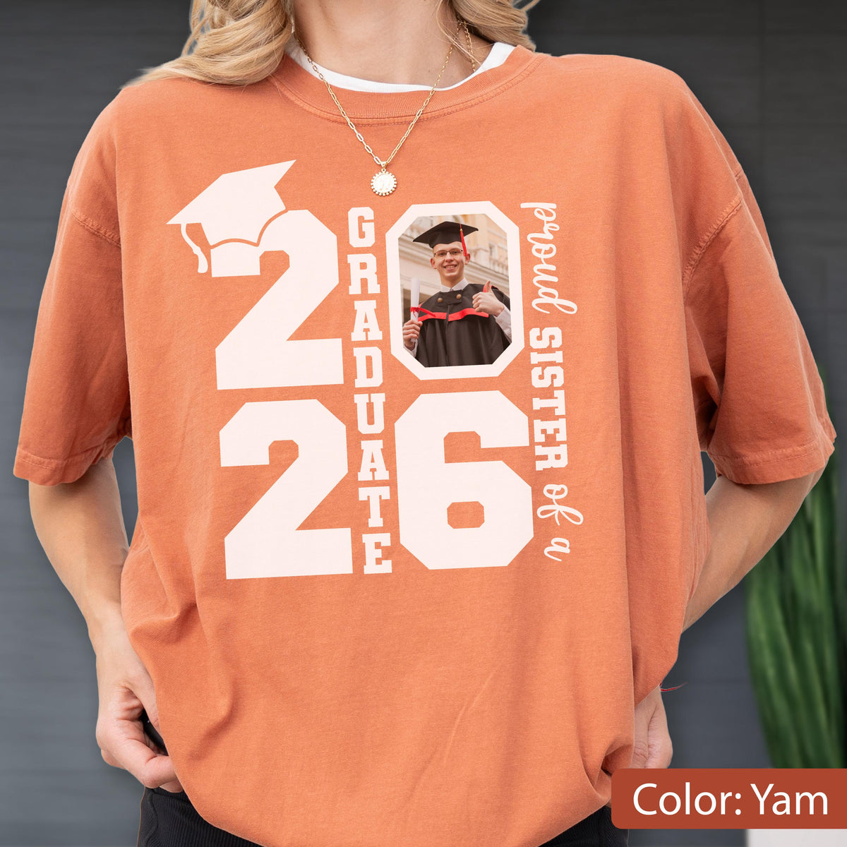 May include: A yam-colored t-shirt with the text "2026 GRADUATE" and a photo of a graduate. The words "proud sister of a" are printed vertically on the right side. A graduation cap graphic is above the number 2026.