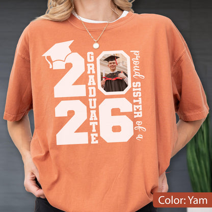May include: A yam-colored t-shirt with the text "2026 GRADUATE" and a photo of a graduate. The words "proud sister of a" are printed vertically on the right side. A graduation cap graphic is above the number 2026.