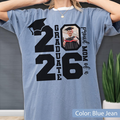May include: Blue jean colored t-shirt with a large black graphic that reads "2026 Graduate". The number "26" has a photo of a graduate in a cap and gown. The text "proud MOM of a" is printed on the right side.