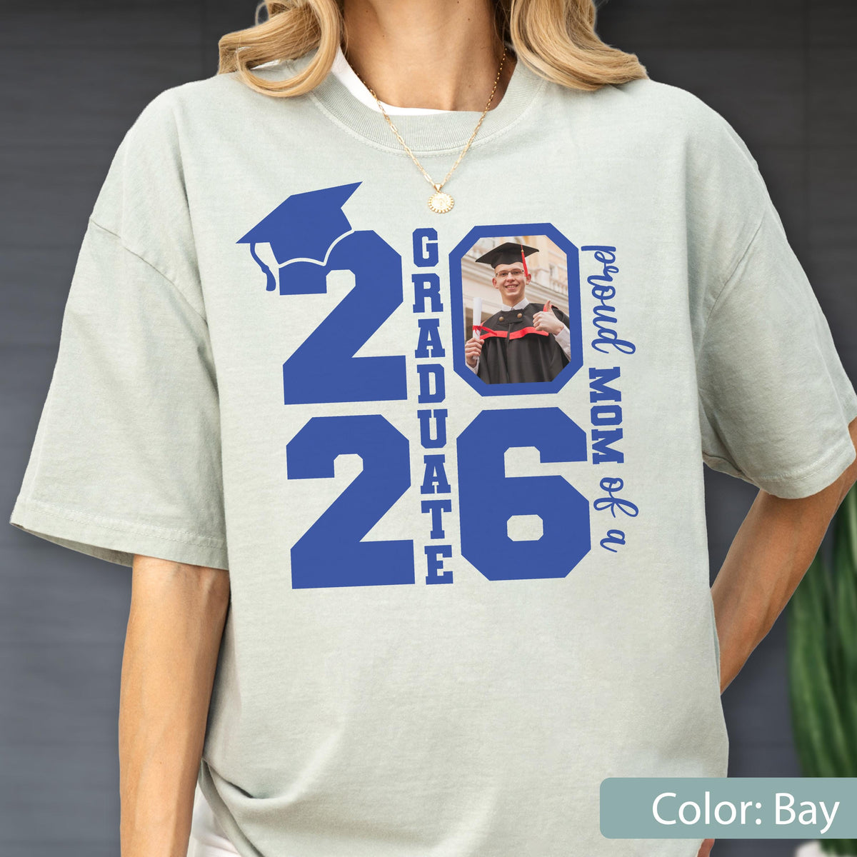 May include: A light bay-colored t-shirt with the text "2026 Graduate" in blue, along with a photo of a graduate. The words "Proud Mom of a" are also printed on the shirt. The shirt has short sleeves.