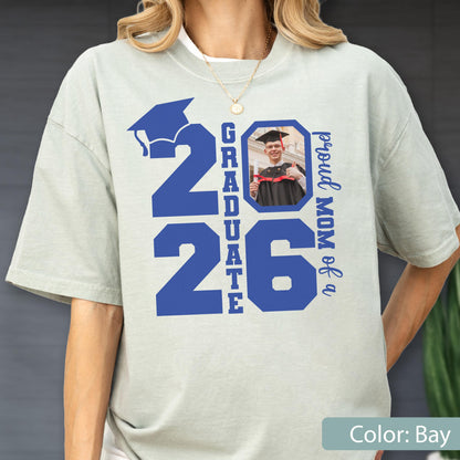 May include: A light bay-colored t-shirt with the text "2026 Graduate" in blue, along with a photo of a graduate. The words "Proud Mom of a" are also printed on the shirt. The shirt has short sleeves.