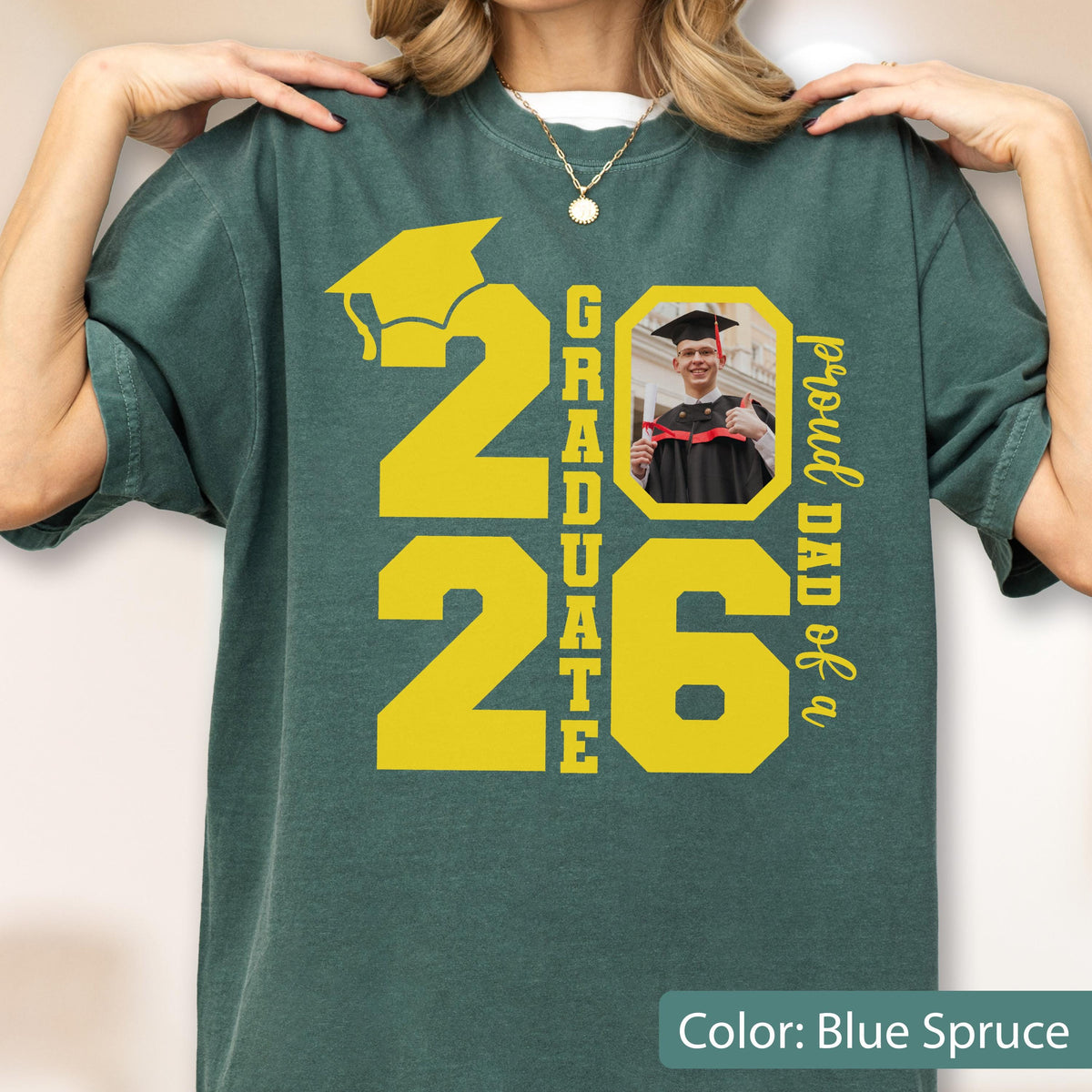 May include: Blue spruce t-shirt with a yellow graduation cap and the numbers "2026". The word "GRADUATE" is vertically printed between the numbers. A photo of a graduate is in a yellow frame, with the text "proud DAD of a" on the right.