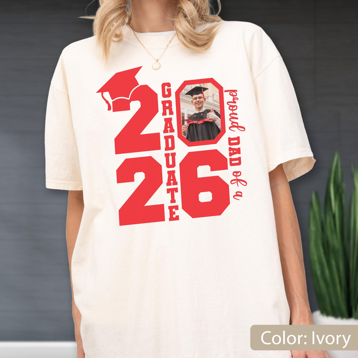 May include: Ivory-colored t-shirt with a red graduation cap graphic and the number "26". The shirt also features the words "GRADUATE" and "proud DAD of a" in red, along with a photo of a graduate.