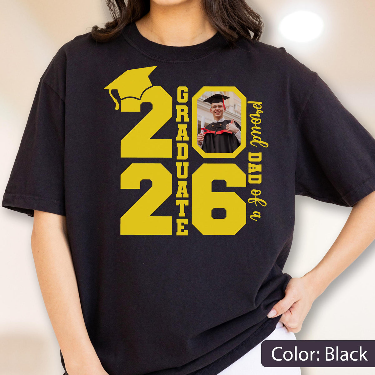 May include: Black t-shirt with a yellow graduation cap graphic and the numbers "2026". The word "GRADUATE" is vertically stacked between the numbers. A photo of a graduate is in the center, with the text "proud DAD of a" on the right.