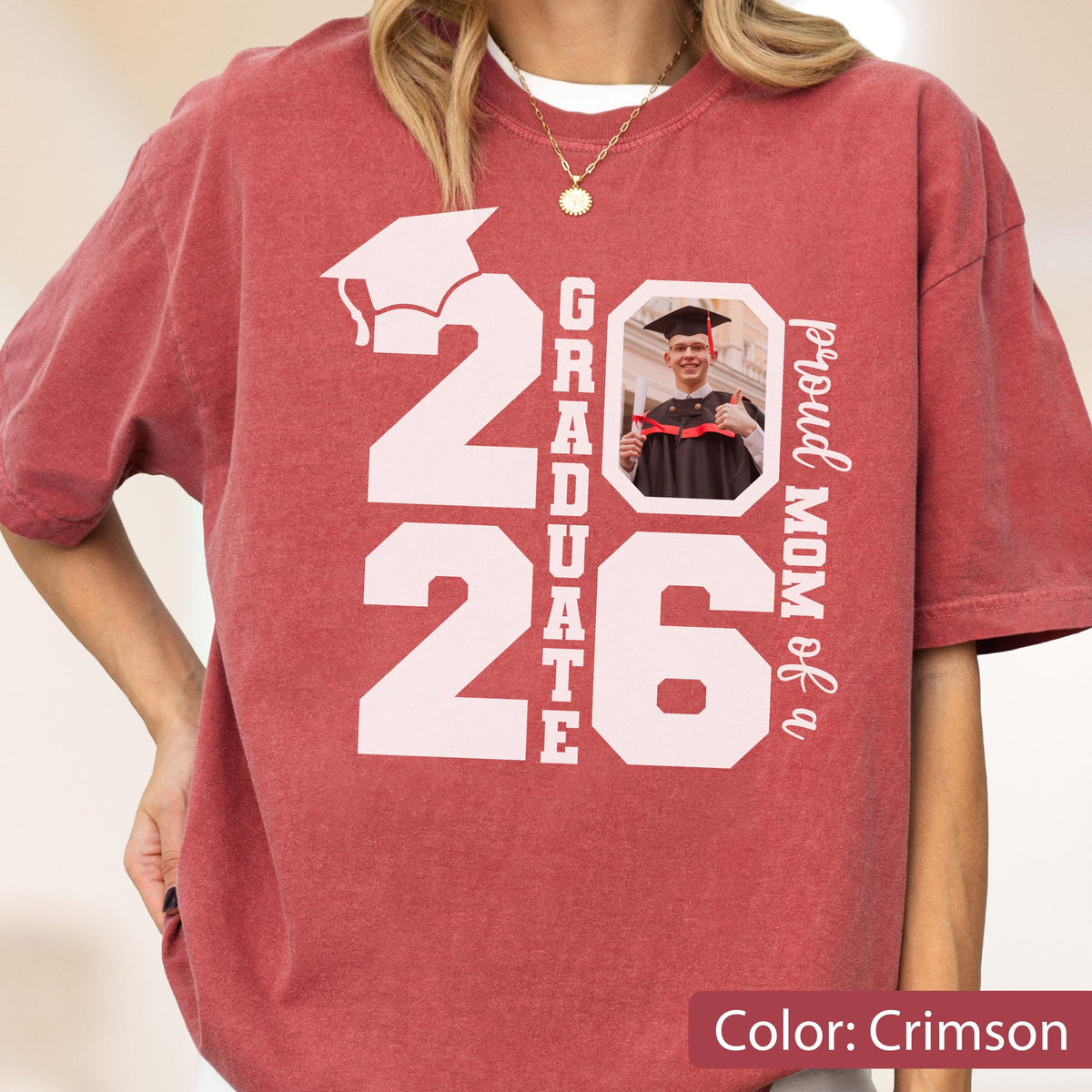 May include: Crimson-colored t-shirt with a large white graphic that reads "2026 GRADUATE" and a photo of a graduate. The words "proud MOM of a" are printed vertically on the right side. The shirt is a comfortable, casual style.