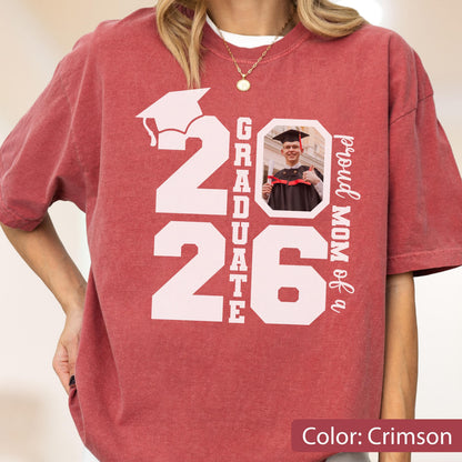 May include: Crimson-colored t-shirt with a large white graphic that reads "2026 GRADUATE" and a photo of a graduate. The words "proud MOM of a" are printed vertically on the right side. The shirt is a comfortable, casual style.