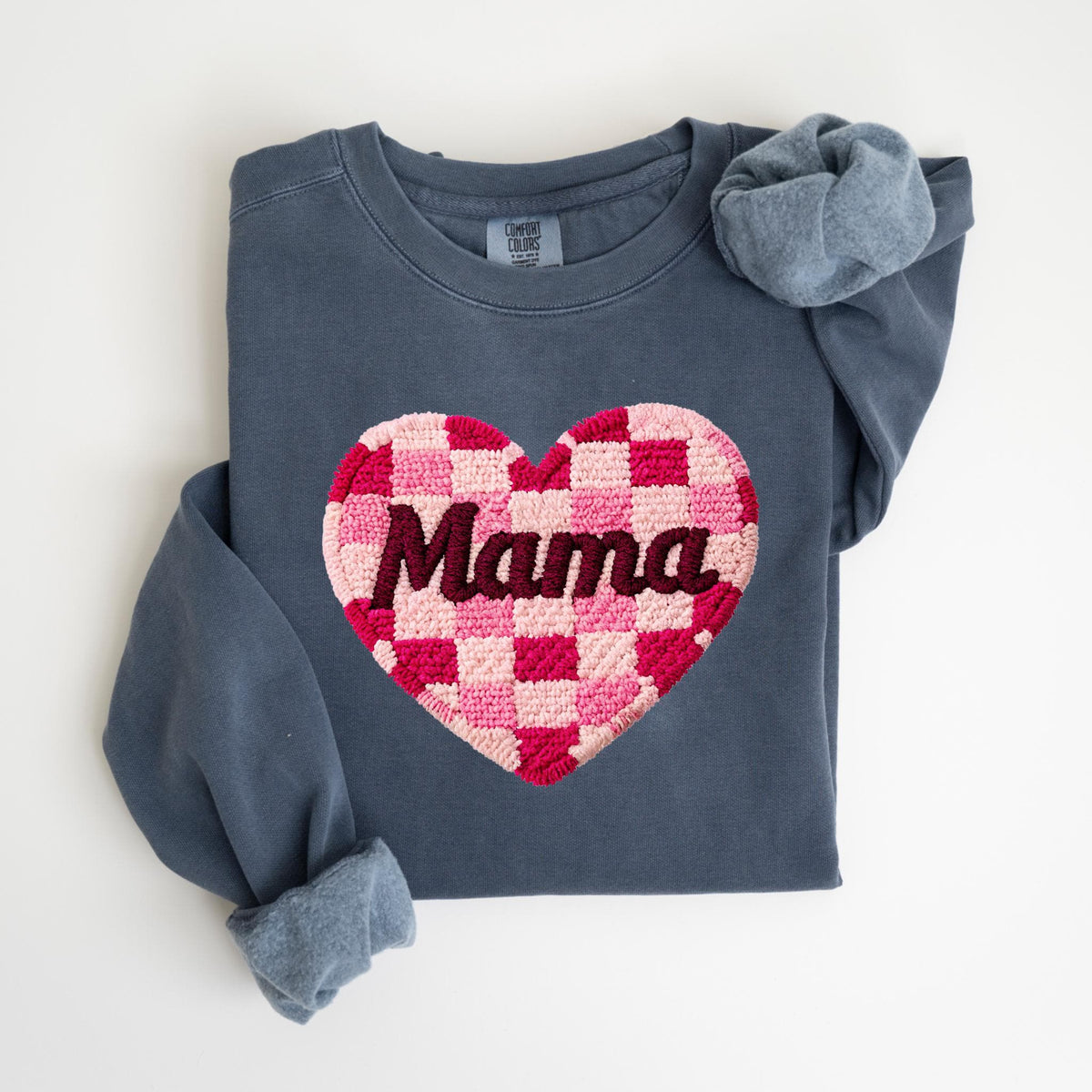 May include: A navy blue sweatshirt with a large, embroidered heart design. The heart is a checkerboard pattern of pink and red squares, with the word "Mama" in dark brown script across the center. A matching scrunchie rests on the sleeve.