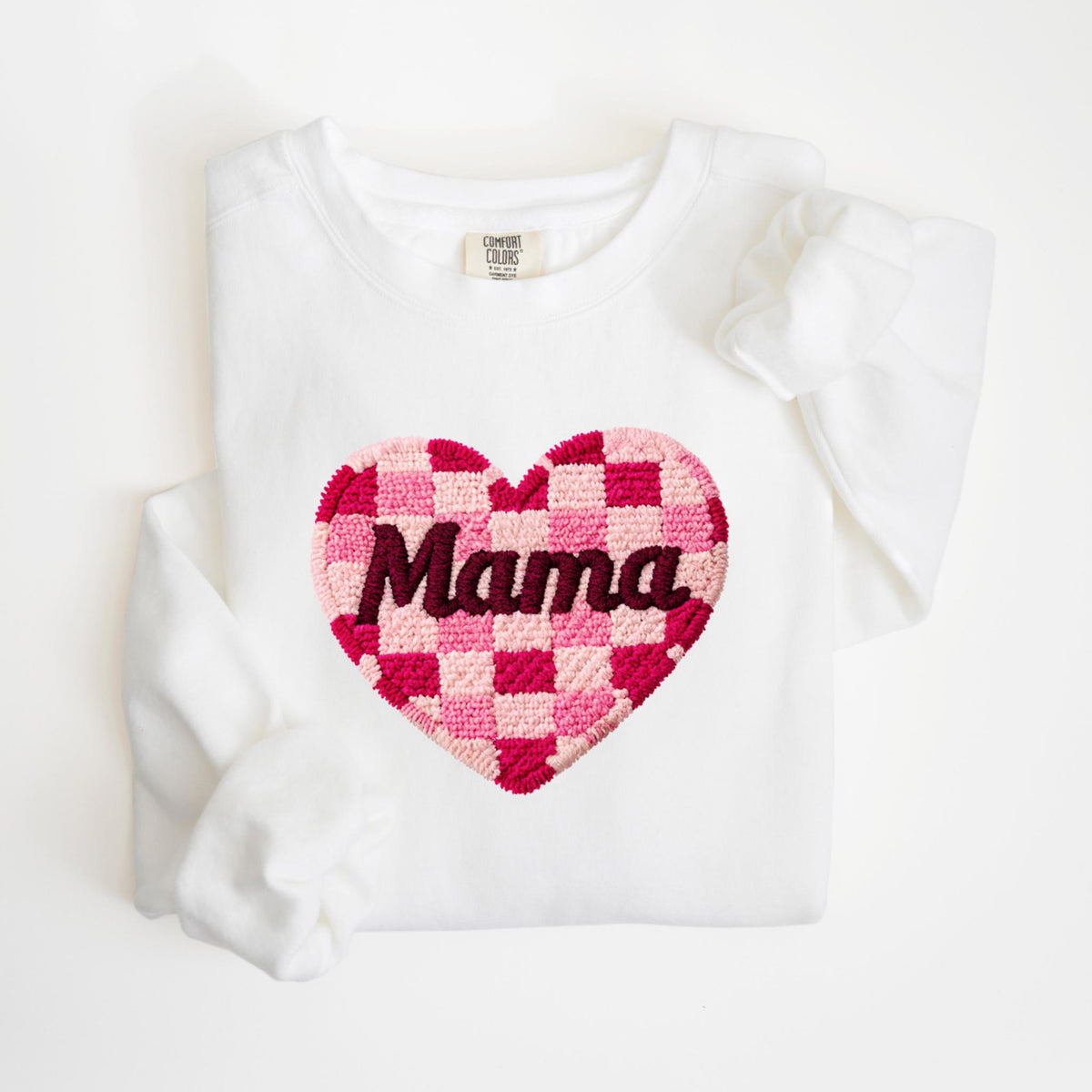 May include: White sweatshirt with a large, heart-shaped design. The heart features a checkerboard pattern in shades of pink and red, with the word "Mama" embroidered in a dark pink color. The sweatshirt has a crew neck and long sleeves.