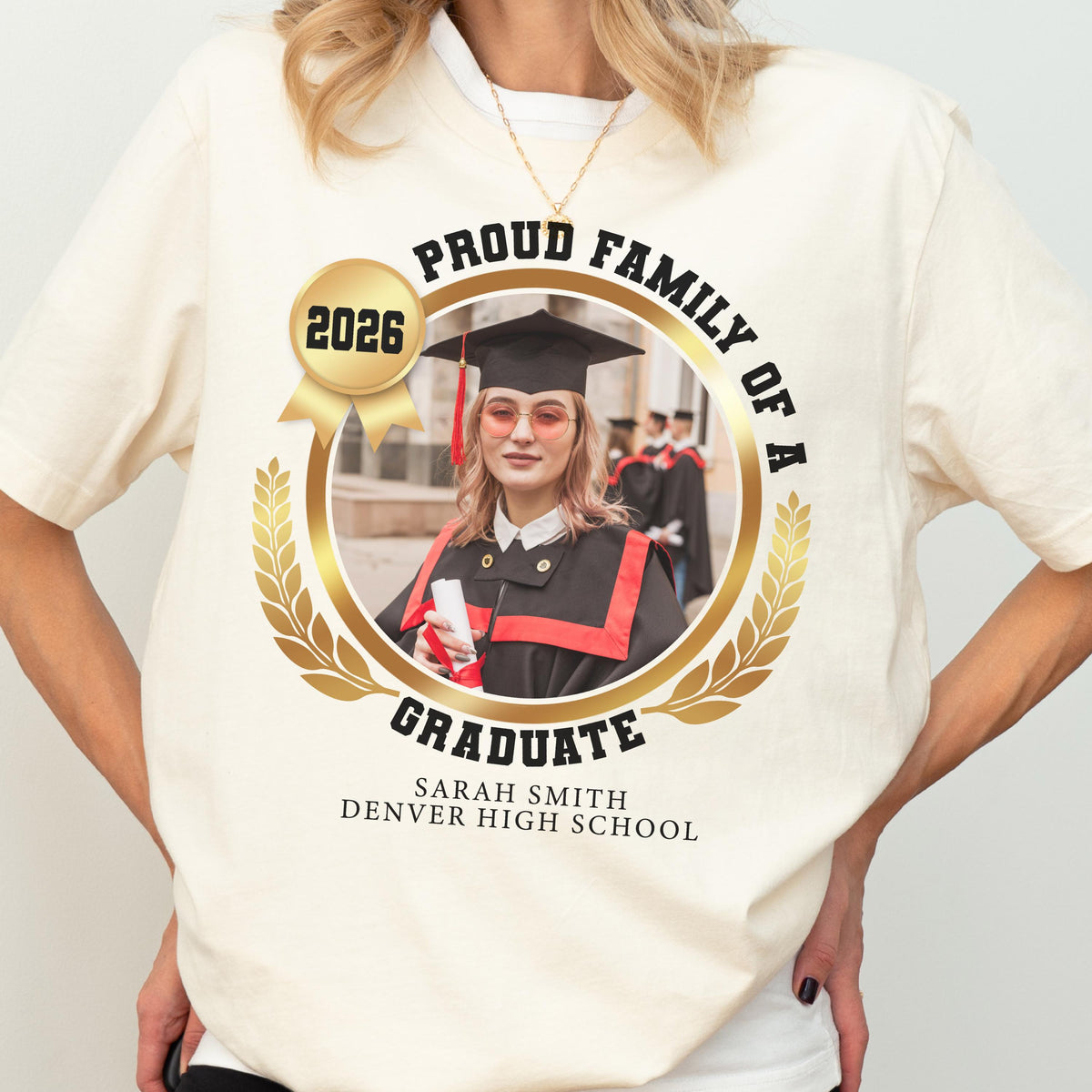 May include: Cream-colored t-shirt with a graduation design. The design features a photo of a graduate in a cap and gown, surrounded by a gold laurel wreath and the text "Proud Family of a Graduate" and "2026". The graduate's name and high school are also printed.