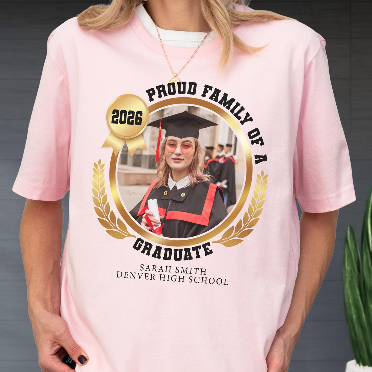 May include: A light pink t-shirt featuring a gold-colored circular design with the words "Proud Family of a Graduate" and the year "2026." The design includes a photo of a graduate in a cap and gown, with the graduate's name and school at the bottom.