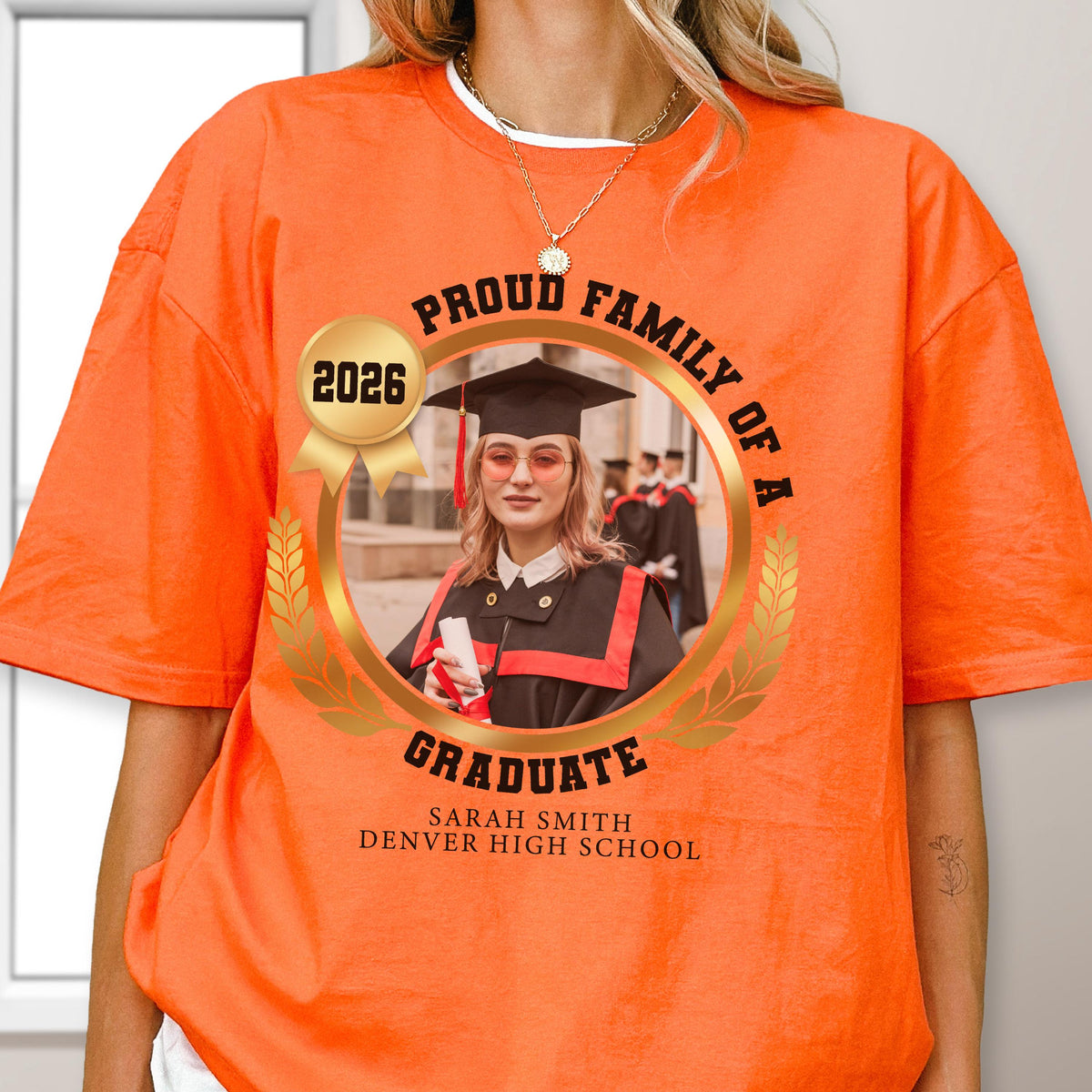 May include: Bright orange t-shirt featuring a graduation design. The design includes a photo of a graduate, the text "Proud Family of a Graduate," the year "2026," and the school name. The shirt is a short-sleeved crew neck.