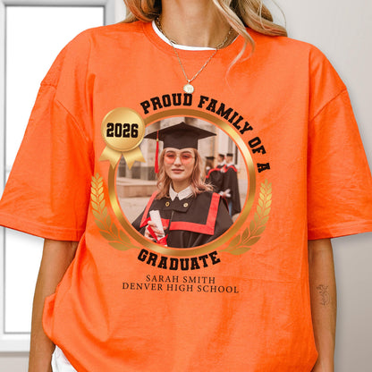 May include: Bright orange t-shirt featuring a graduation design. The design includes a photo of a graduate, the text "Proud Family of a Graduate," the year "2026," and the school name. The shirt is a short-sleeved crew neck.