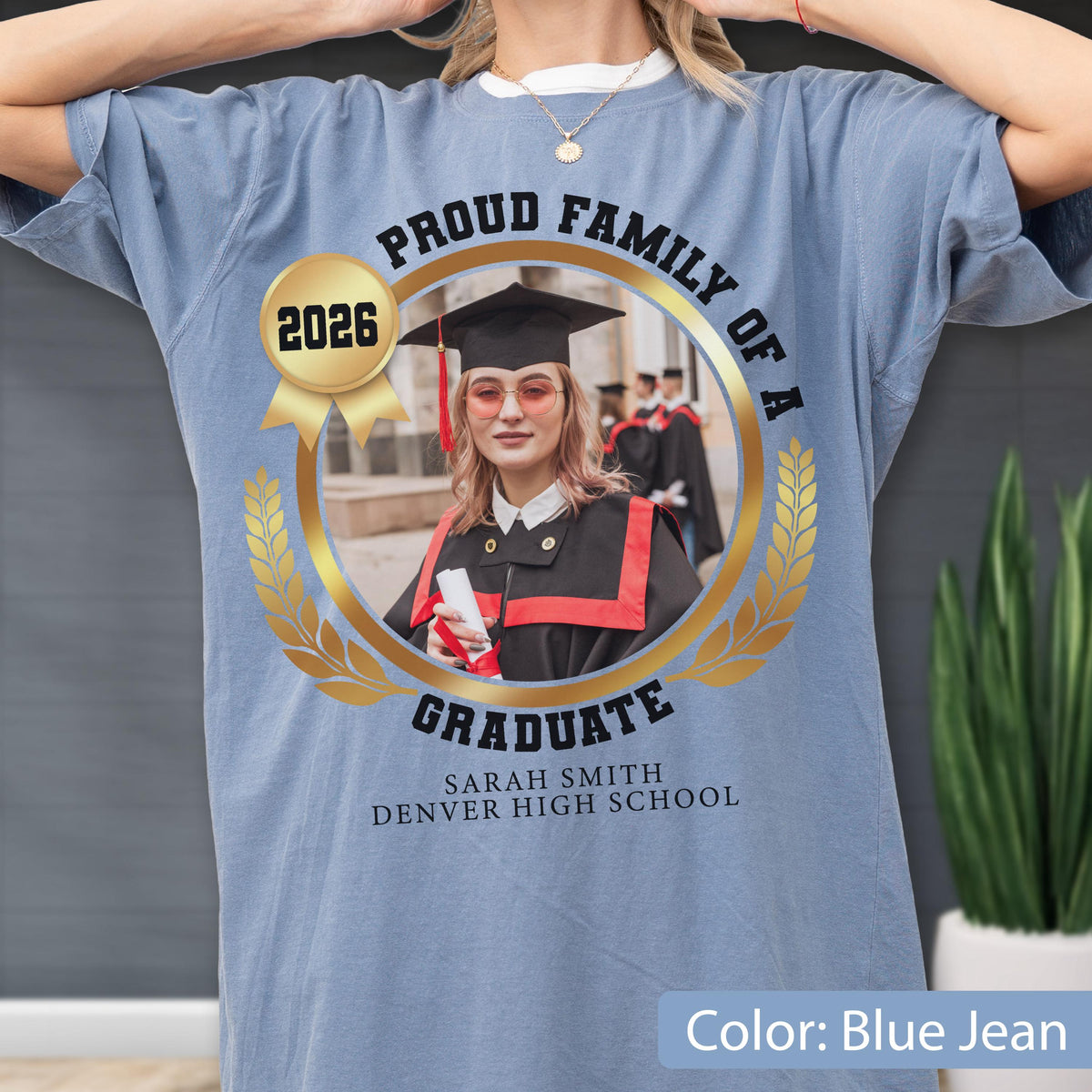 May include: Blue jean colored t-shirt with a gold-framed photo of a graduate wearing a cap and gown. The shirt features the text "PROUD FAMILY OF A GRADUATE" and "2026" with the graduate's name and high school.