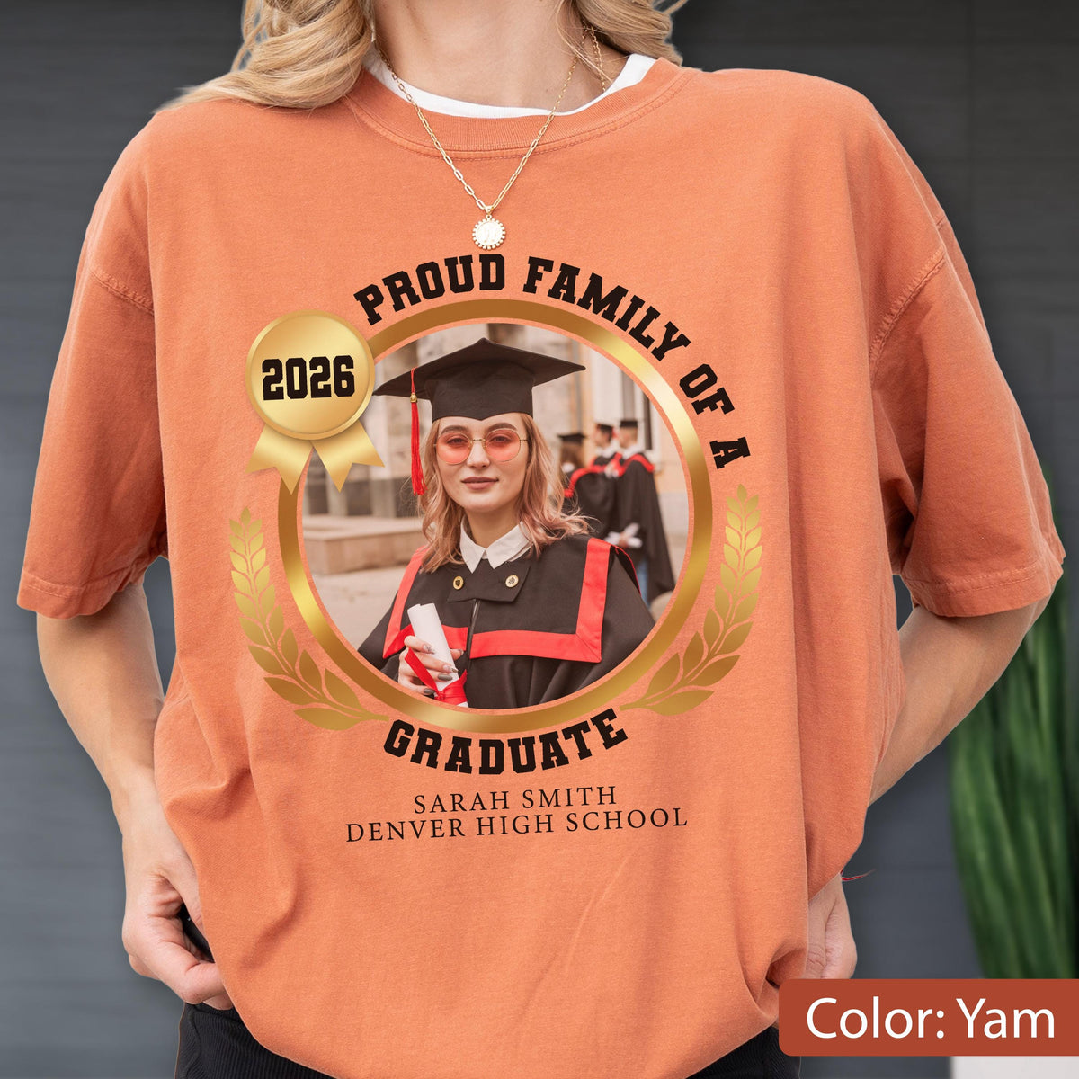 May include: A yam-colored t-shirt featuring a graduation design. The design includes a photo of a graduate in a cap and gown, surrounded by text that reads "Proud Family of a Graduate" and the graduate's name and school.