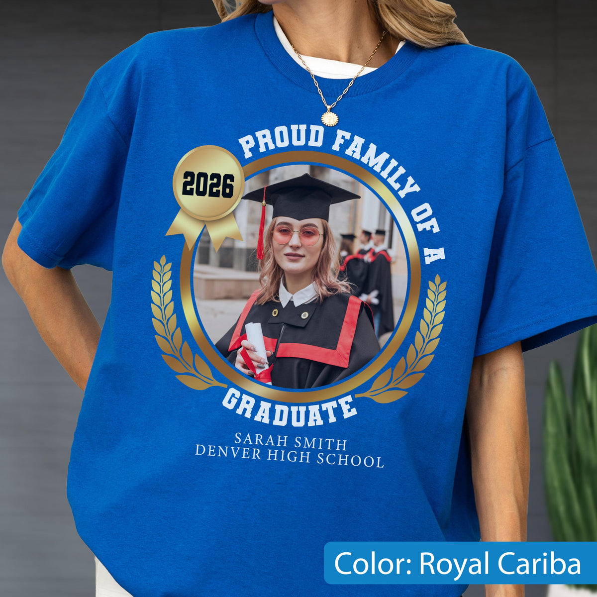 May include: Royal Cariba blue t-shirt with a gold emblem featuring a graduate's photo, the year 2026, and the text "PROUD FAMILY OF A GRADUATE." The graduate's name and high school are also printed on the shirt.