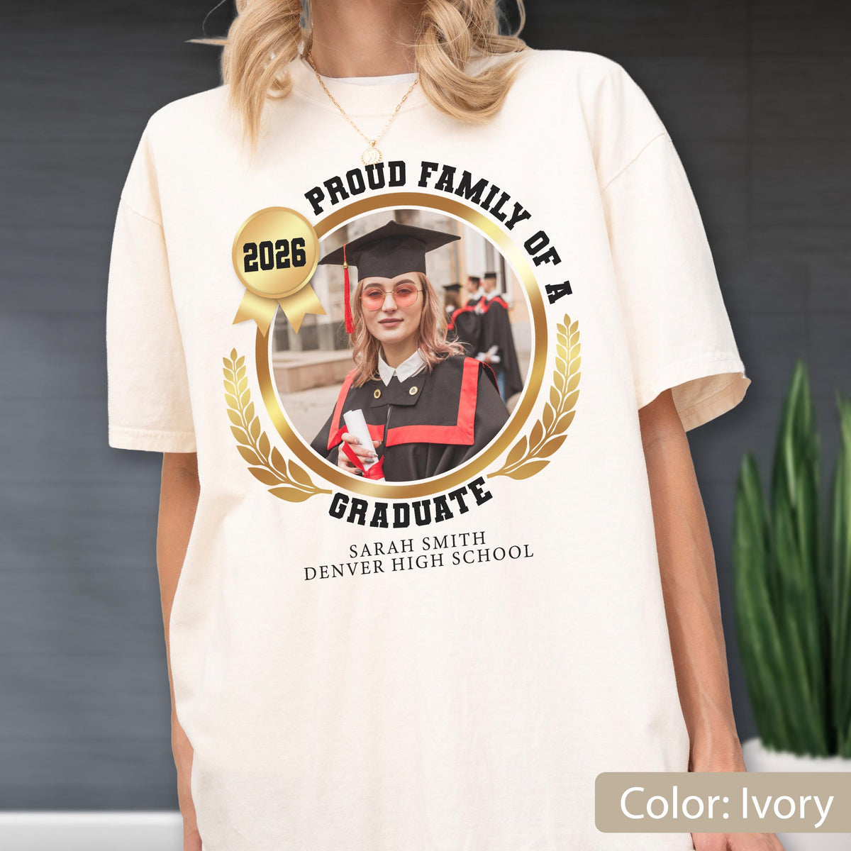 May include: Ivory-colored t-shirt featuring a graduation design. The design includes a photo of a graduate, the text "Proud Family of a Graduate," the year "2026," and the graduate's name and high school. The design is framed by a gold laurel wreath.