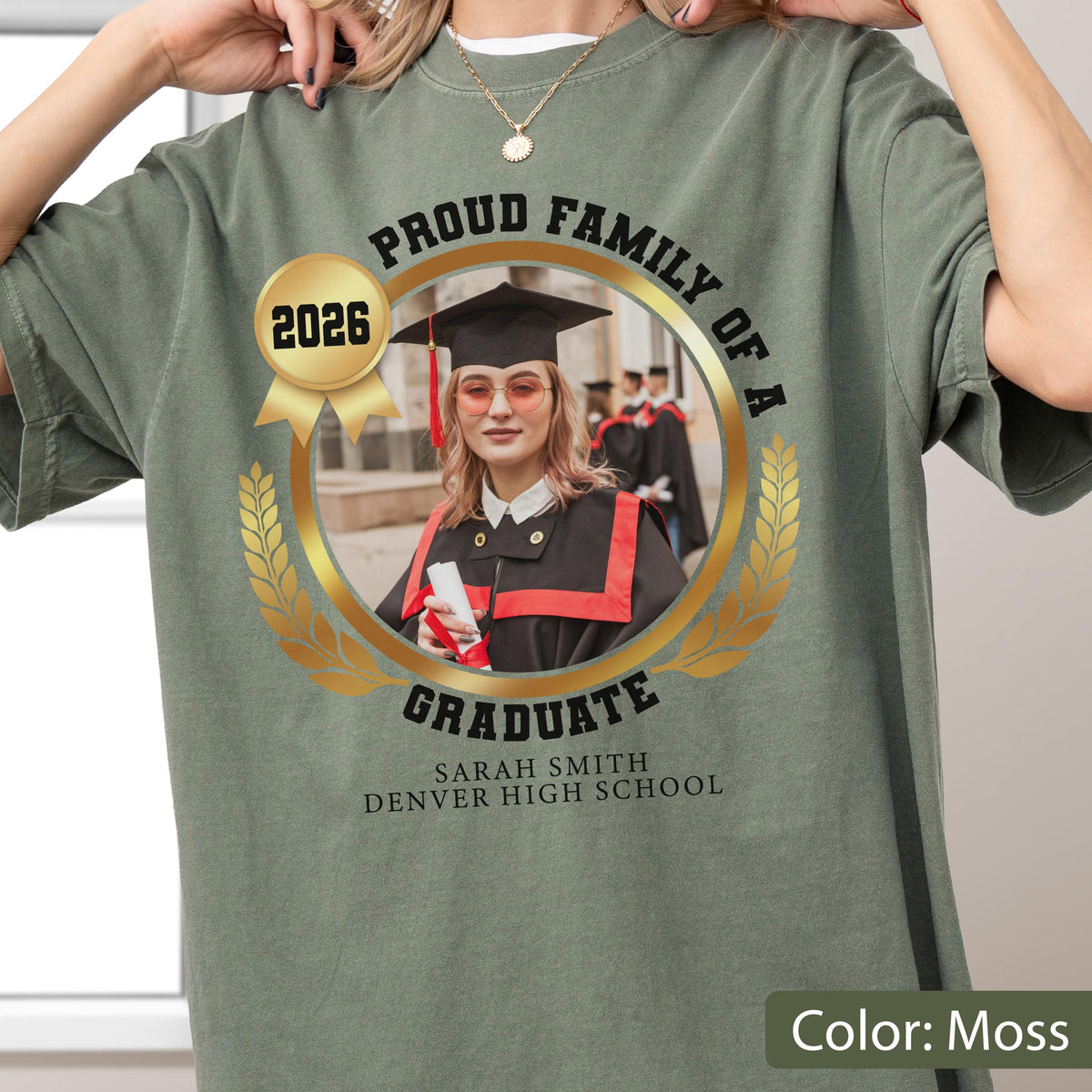 May include: Moss green t-shirt with a gold emblem featuring a graduate in a cap and gown. The emblem reads "Proud Family of a Graduate" and includes the year "2026", the graduate's name, and high school. The shirt's color is labeled "Color: Moss".