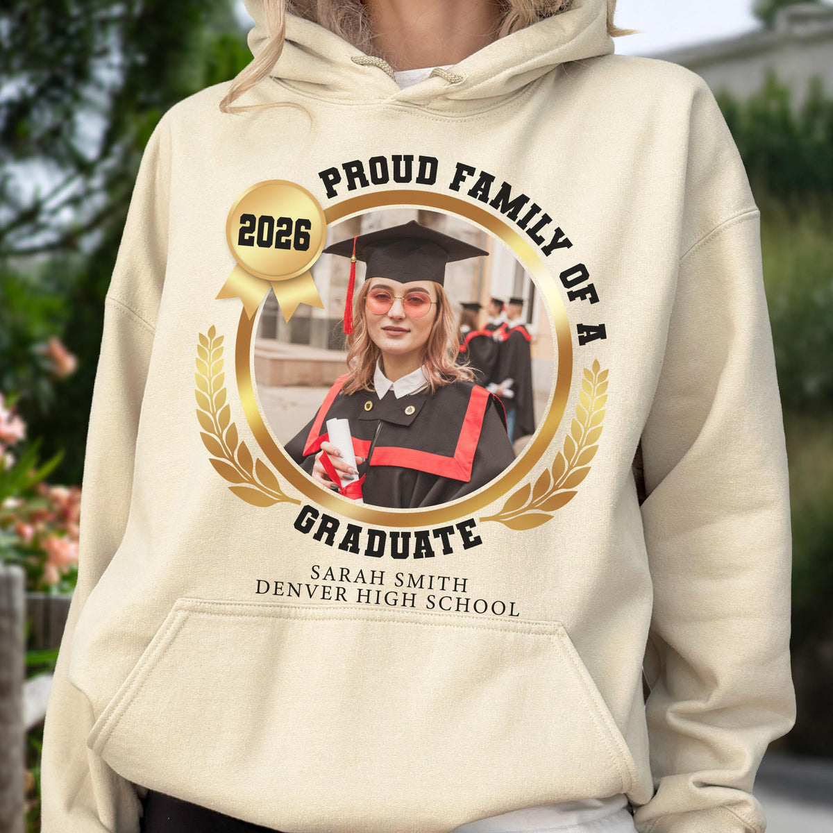 May include: A beige hoodie featuring a gold emblem with the text "PROUD FAMILY OF A GRADUATE" and "2026." The emblem surrounds a photo of a graduate in a cap and gown. The text "SARAH SMITH DENVER HIGH SCHOOL" is below the emblem.