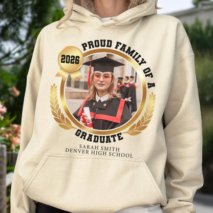 May include: A beige hoodie featuring a gold emblem with the text "PROUD FAMILY OF A GRADUATE" and "2026." The emblem surrounds a photo of a graduate in a cap and gown. The text "SARAH SMITH DENVER HIGH SCHOOL" is below the emblem.