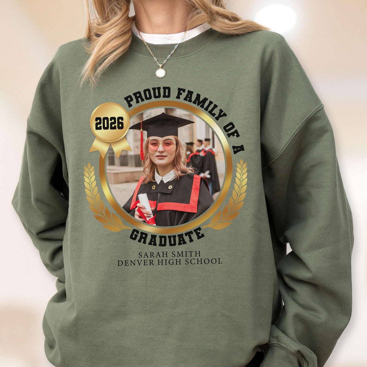 May include: A sage green sweatshirt featuring a gold emblem with the text "Proud Family of a Graduate" and the year "2026." The emblem includes a photo of a graduate in a cap and gown, with the graduate's name and high school below.