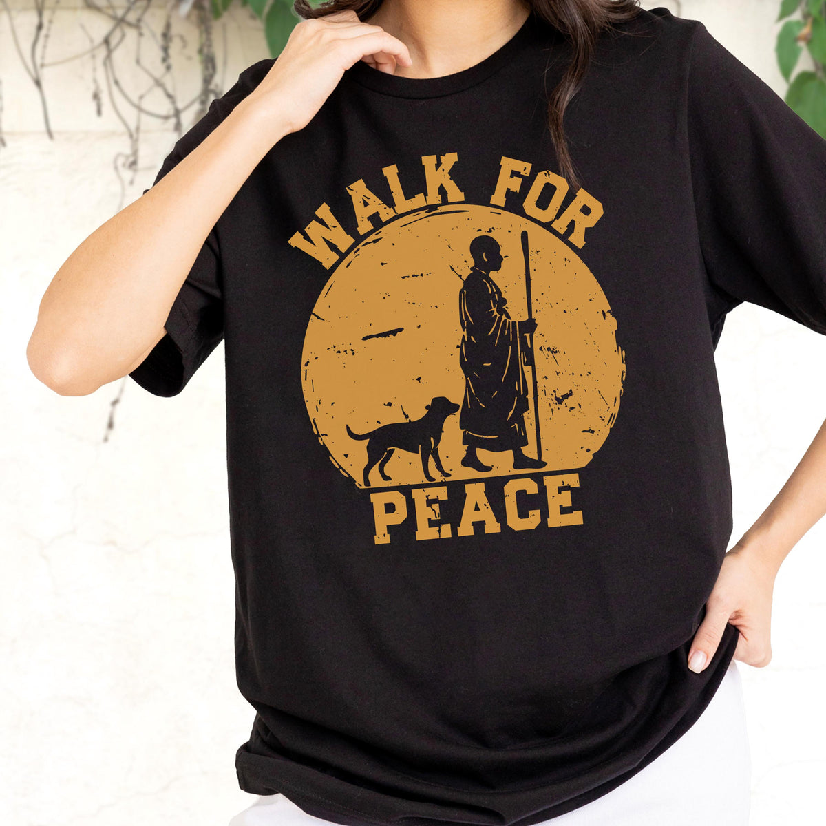 May include: A black short-sleeved t-shirt featuring a large circular mustard-yellow graphic on the chest. The design depicts a silhouette of a person in robes holding a staff walking with a dog, surrounded by the text "WALK FOR PEACE" in bold letters.