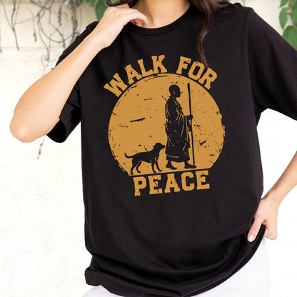 May include: A black short-sleeved t-shirt featuring a large circular mustard-yellow graphic on the chest. The design depicts a silhouette of a person in robes holding a staff walking with a dog, surrounded by the text "WALK FOR PEACE" in bold letters.
