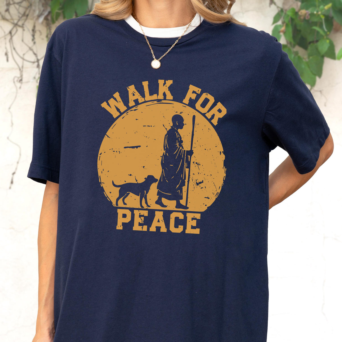 May include: A navy blue short-sleeved t-shirt featuring a graphic of a person in robes walking with a dog and a staff inside a large mustard yellow circle. The text "WALK FOR PEACE" is printed in mustard yellow block letters above and below the circle.