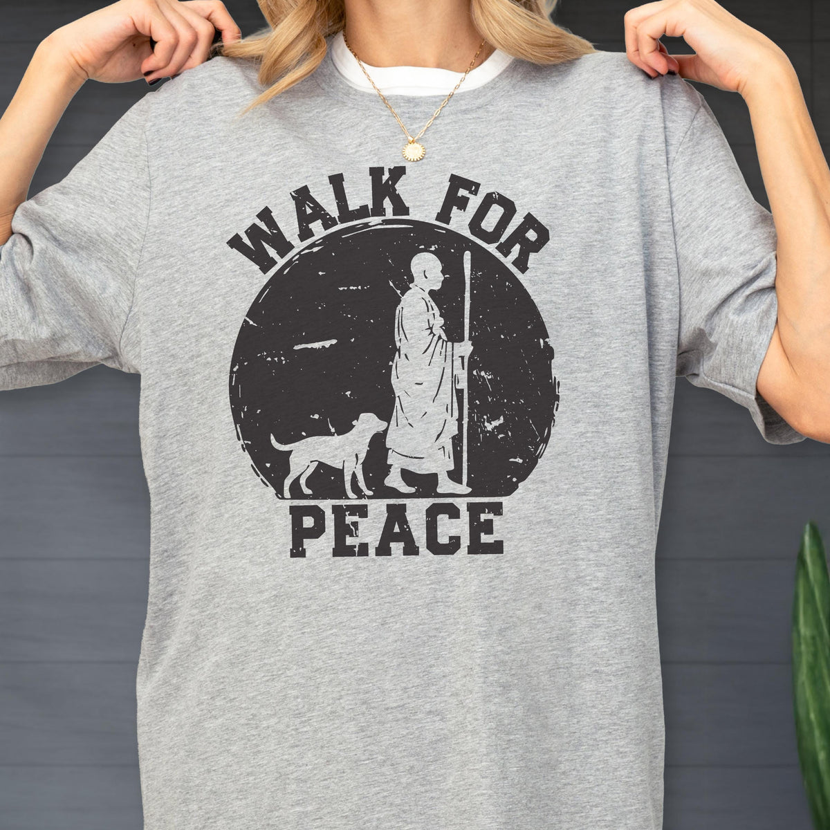 May include: A heather gray short-sleeve t-shirt featuring a black graphic print of a person in robes walking with a staff alongside a dog. The design is set within a distressed circle and includes the text WALK FOR PEACE in bold black lettering.