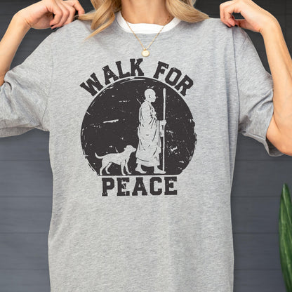 May include: A heather gray short-sleeve t-shirt featuring a black graphic print of a person in robes walking with a staff alongside a dog. The design is set within a distressed circle and includes the text WALK FOR PEACE in bold black lettering.