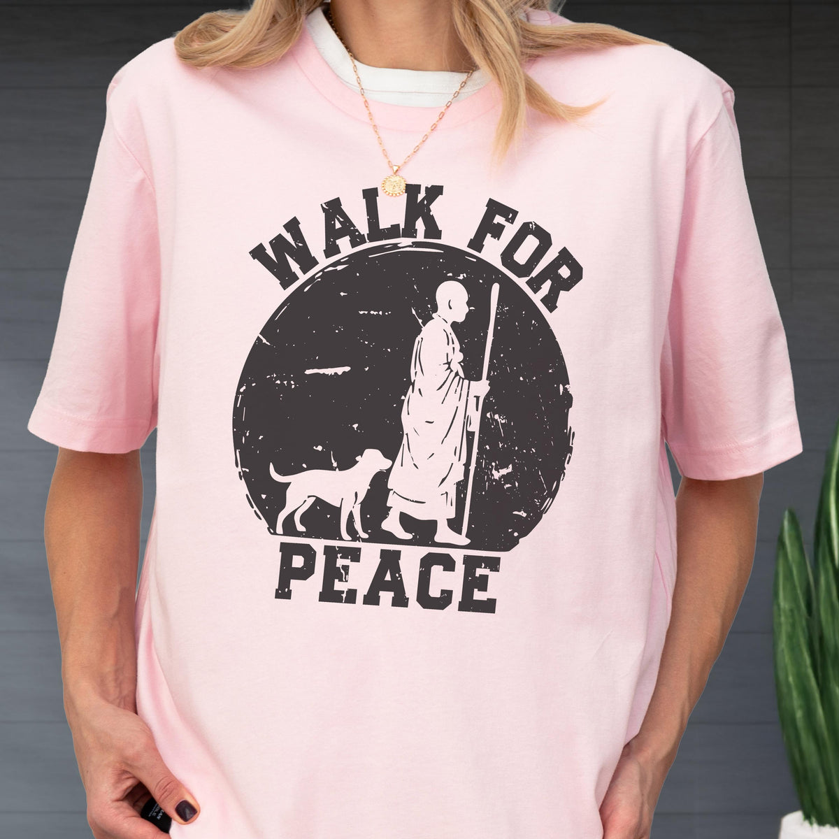 May include: A light pink short-sleeved t-shirt featuring a black circular graphic. The design depicts a person in robes walking with a staff alongside a dog, with the text "WALK FOR PEACE" printed in bold, distressed black capital letters.