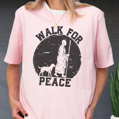 May include: A light pink short-sleeved t-shirt featuring a black circular graphic. The design depicts a person in robes walking with a staff alongside a dog, with the text "WALK FOR PEACE" printed in bold, distressed black capital letters.
