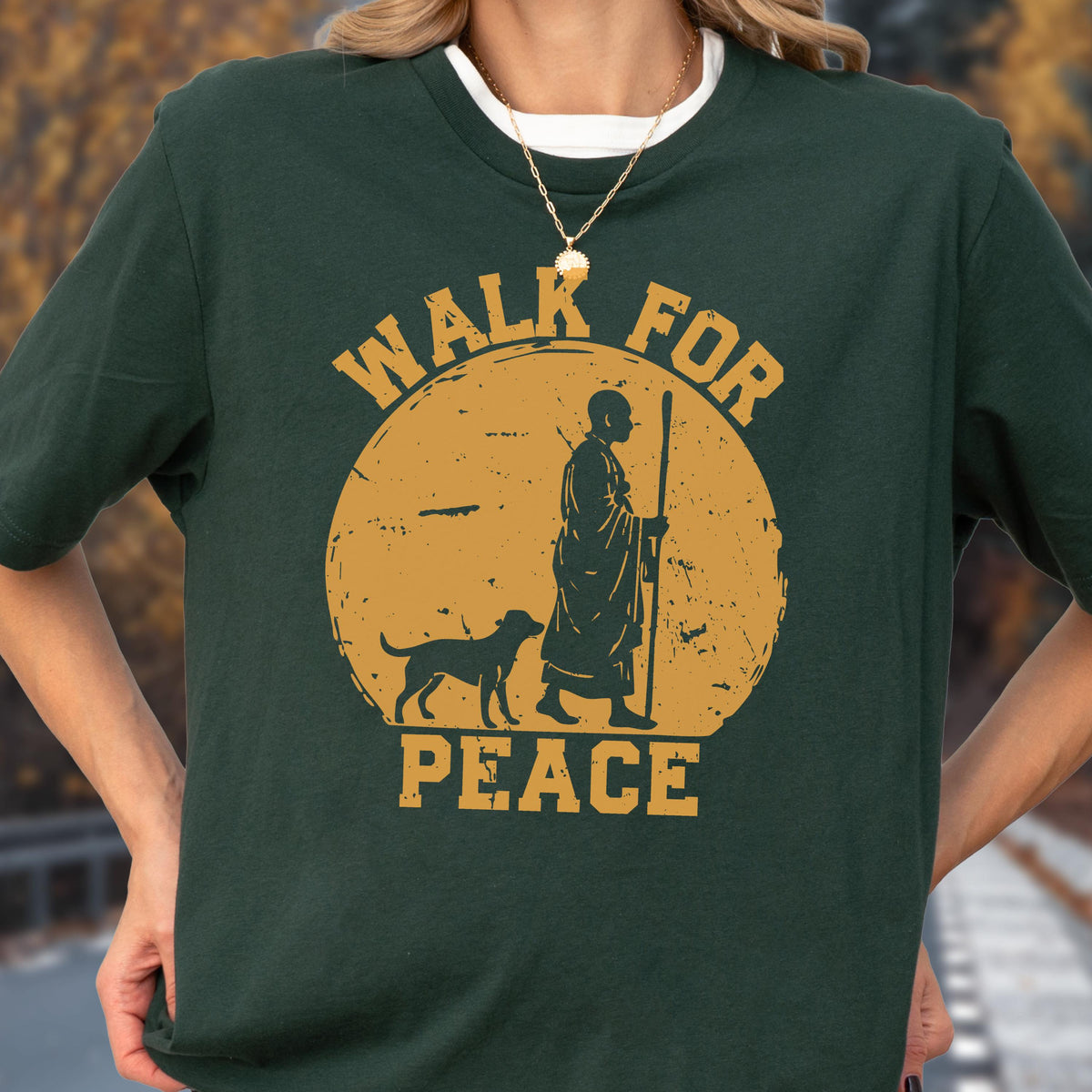 May include: A dark green t-shirt featuring a gold graphic of a monk walking with a dog in front of a large circle. The text "WALK FOR PEACE" is printed in gold capital letters above and below the circular graphic. The shirt has a crew neckline.