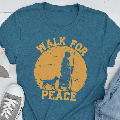 May include: A teal short-sleeved t-shirt featuring a distressed yellow circular graphic. The design depicts a person in robes walking with a staff alongside a dog, with the text WALK FOR PEACE arched around the circle in a bold, serif font.