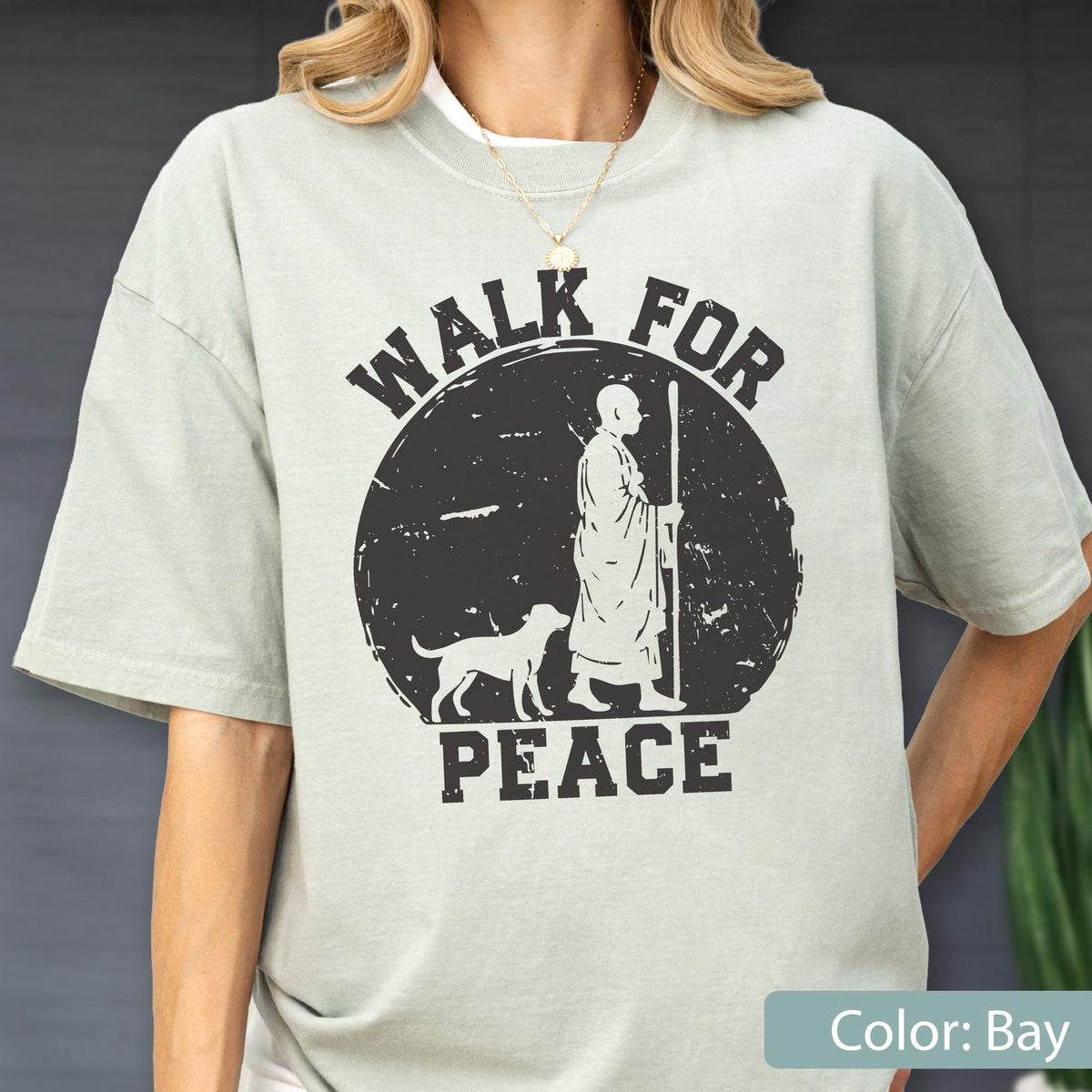 May include: A light green t-shirt with a black graphic that reads "WALK FOR PEACE." The graphic depicts a person with a dog and staff inside a circle. The shirt is a solid color and has short sleeves.