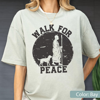 May include: A light green t-shirt with a black graphic that reads "WALK FOR PEACE." The graphic depicts a person with a dog and staff inside a circle. The shirt is a solid color and has short sleeves.