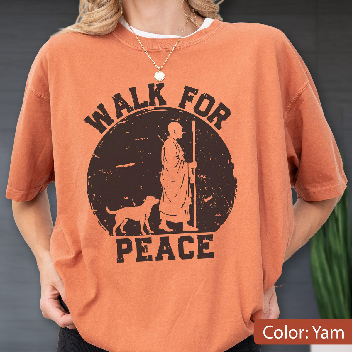 May include: Orange-colored t-shirt with a graphic design. The design features a figure with a dog, a staff, and the words "WALK FOR PEACE" in a circular arrangement. The shirt is a solid color and has short sleeves.