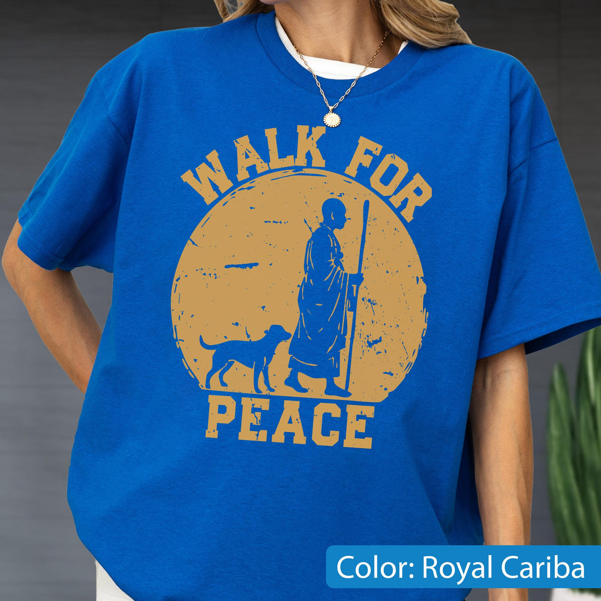 May include: Royal Cariba blue t-shirt with a round, distressed gold graphic. The graphic features a figure with a staff, a dog, and the words "WALK FOR PEACE". The shirt has short sleeves and a crew neck.