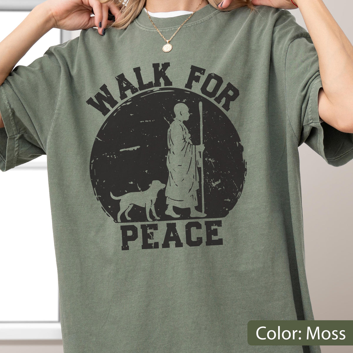 May include: Moss green t-shirt with a black graphic. The graphic features a figure with a dog and the words "WALK FOR PEACE" in a circular design. The shirt has short sleeves and a crew neck.