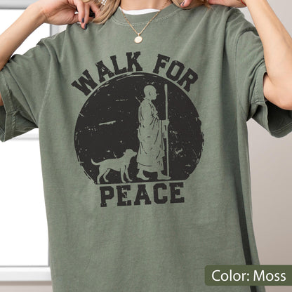 May include: Moss green t-shirt with a black graphic. The graphic features a figure with a dog and the words "WALK FOR PEACE" in a circular design. The shirt has short sleeves and a crew neck.