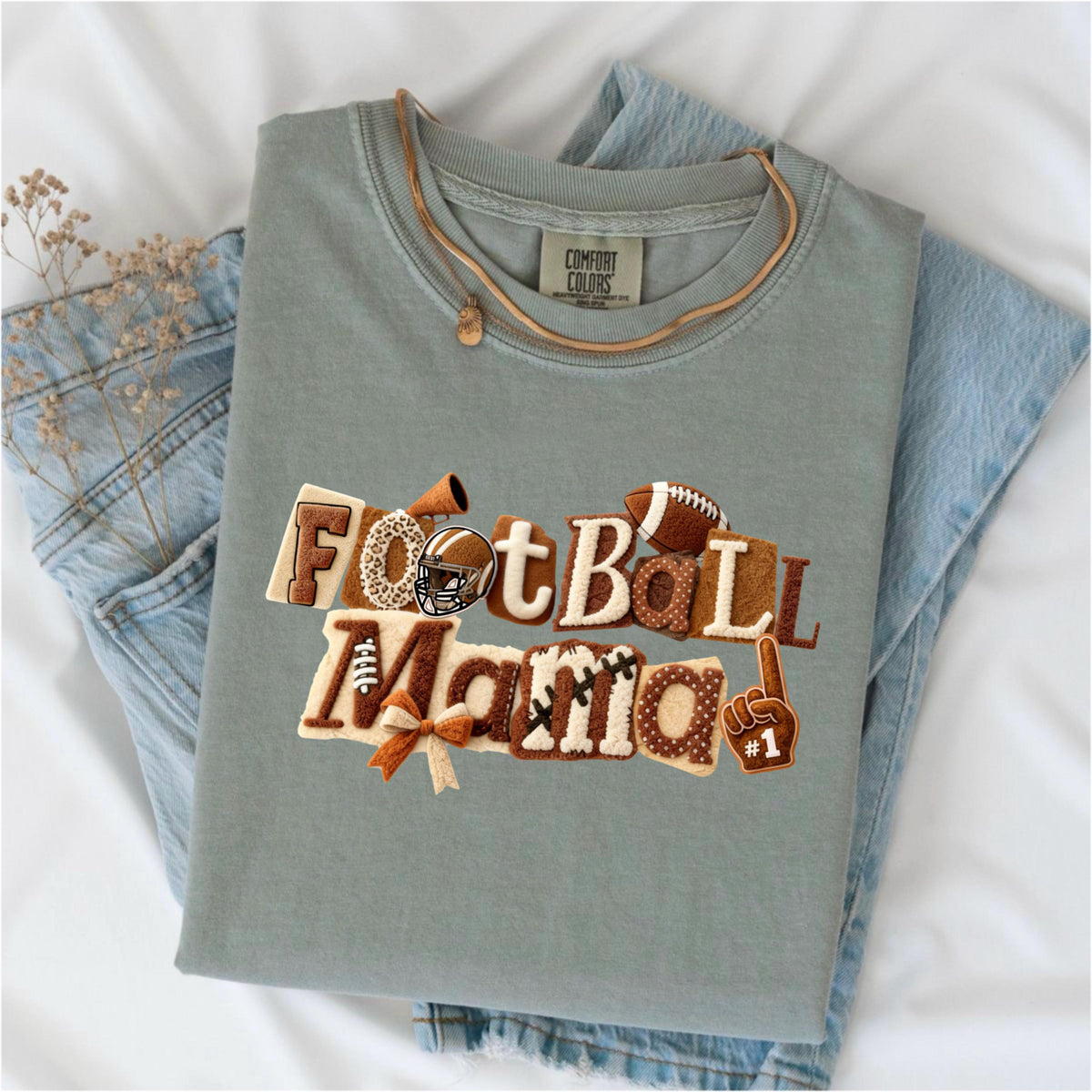 May include: A sage green t-shirt with the words "FootBall Mama" in a decorative, textured design. The design includes footballs, a helmet, and a #1 foam finger. The shirt is folded on top of a pair of blue jeans.