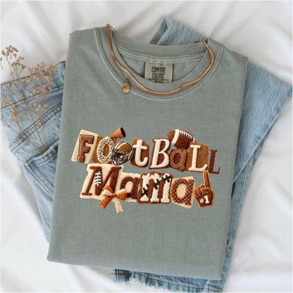 May include: A sage green t-shirt with the words "FootBall Mama" in a decorative, textured design. The design includes footballs, a helmet, and a #1 foam finger. The shirt is folded on top of a pair of blue jeans.