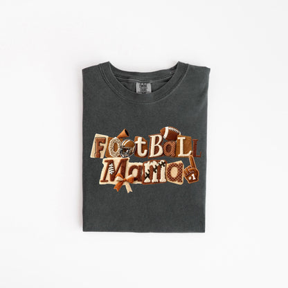 May include: Dark grey t-shirt with the words "Football Mama" in a decorative, layered design. The design includes footballs, a helmet, a bow, and a foam finger. The shirt is folded neatly on a white surface.