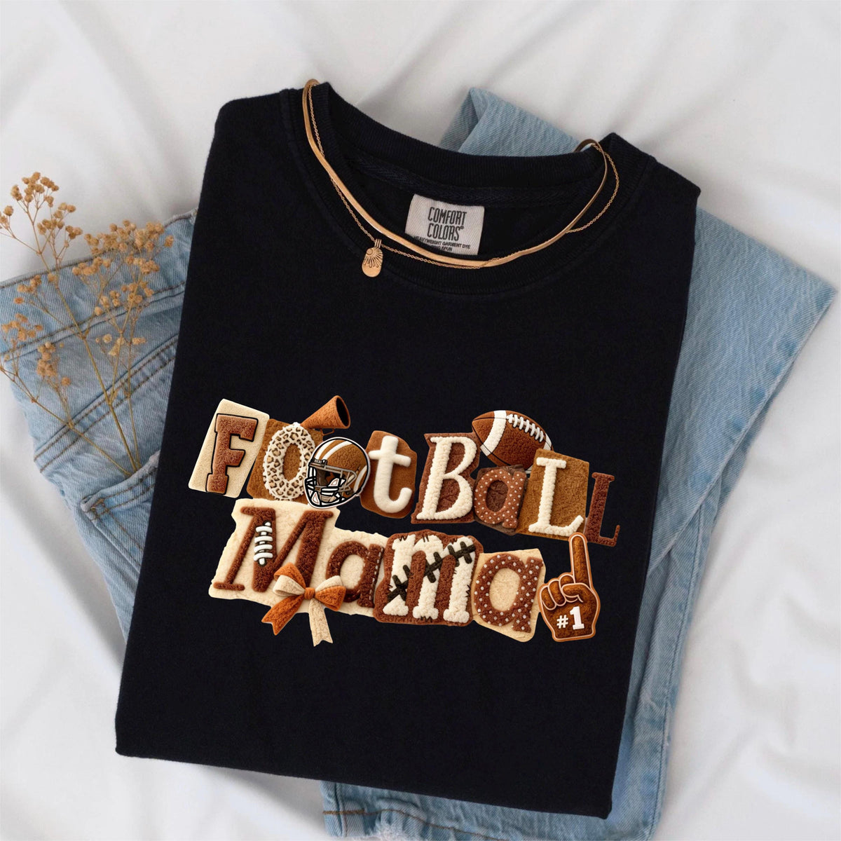 May include: Black t-shirt with the embroidered text "Football Mama" in brown and cream colors, featuring football-themed graphics. A gold necklace and blue jeans are also visible.