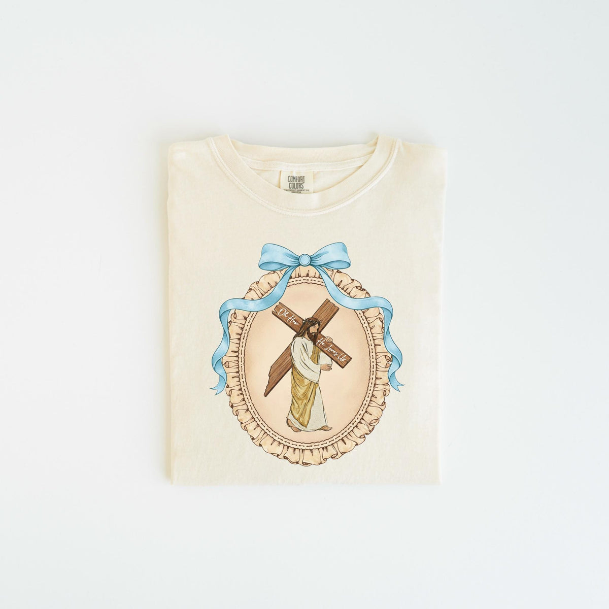 May include: Cream-colored t-shirt featuring a religious illustration. The design includes a depiction of Jesus carrying a cross, framed by a blue ribbon and an oval border. The text on the cross is visible. The shirt is folded neatly.