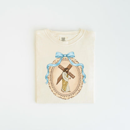 May include: Cream-colored t-shirt featuring a religious illustration. The design includes a depiction of Jesus carrying a cross, framed by a blue ribbon and an oval border. The text on the cross is visible. The shirt is folded neatly.