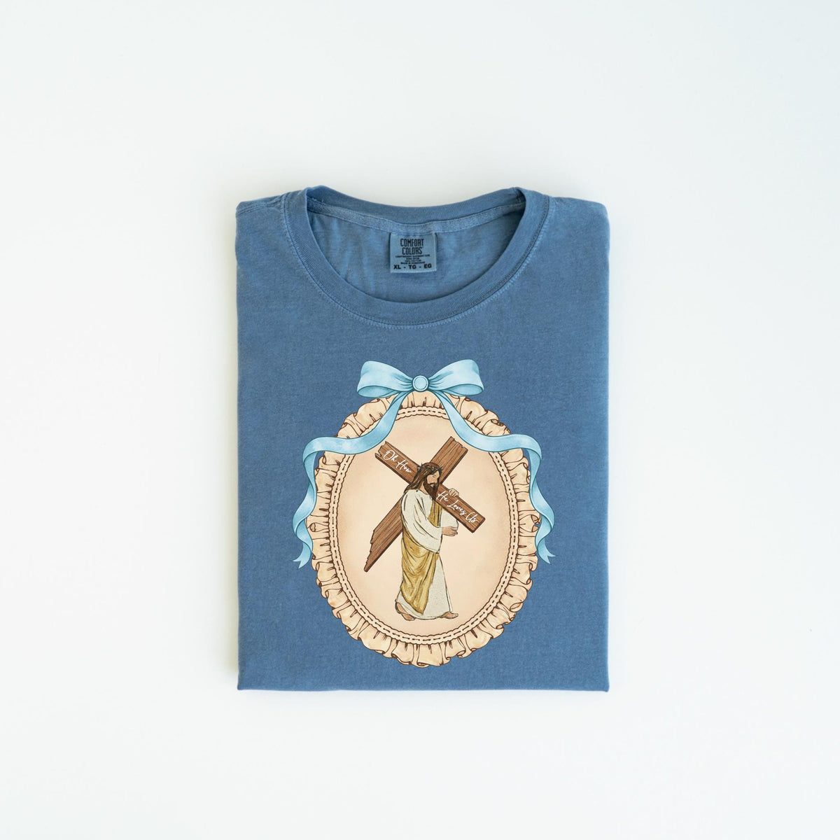 May include: Blue t-shirt featuring a framed illustration of Jesus carrying a cross. The illustration is surrounded by a light blue ribbon and bow. The text on the cross reads "For Him".