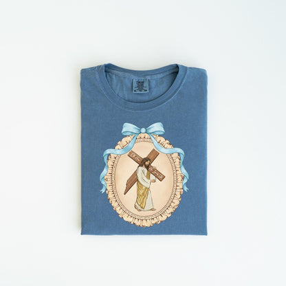 May include: Blue t-shirt featuring a framed illustration of Jesus carrying a cross. The illustration is surrounded by a light blue ribbon and bow. The text on the cross reads "For Him".